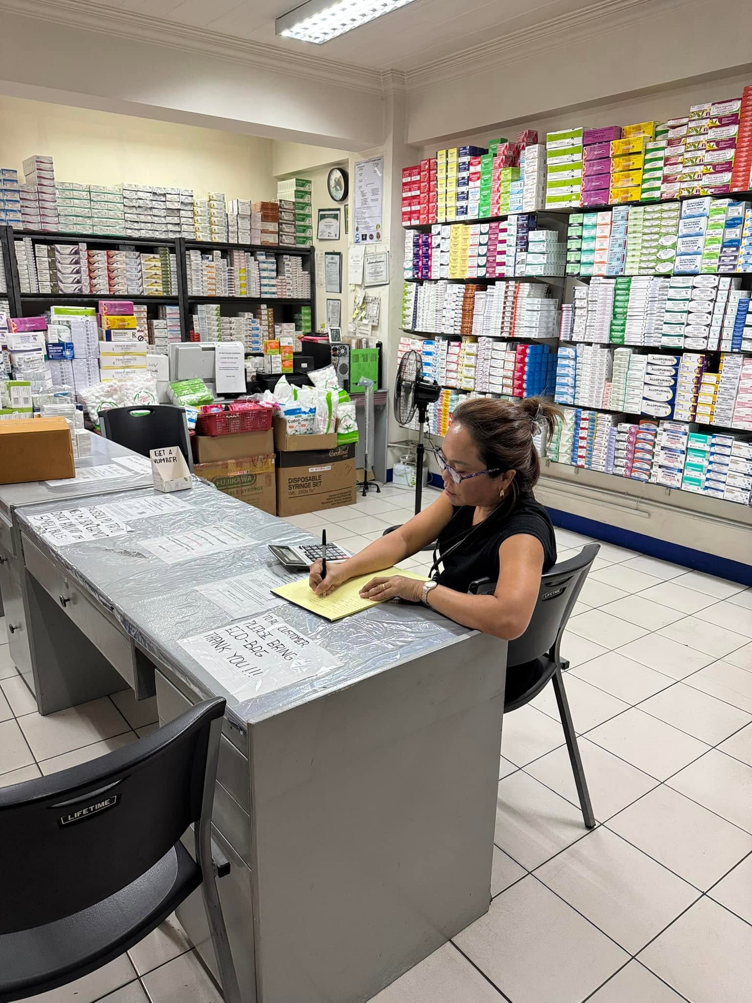A woman writes at a pharmacy counter surrounded by shelves of medicines.
